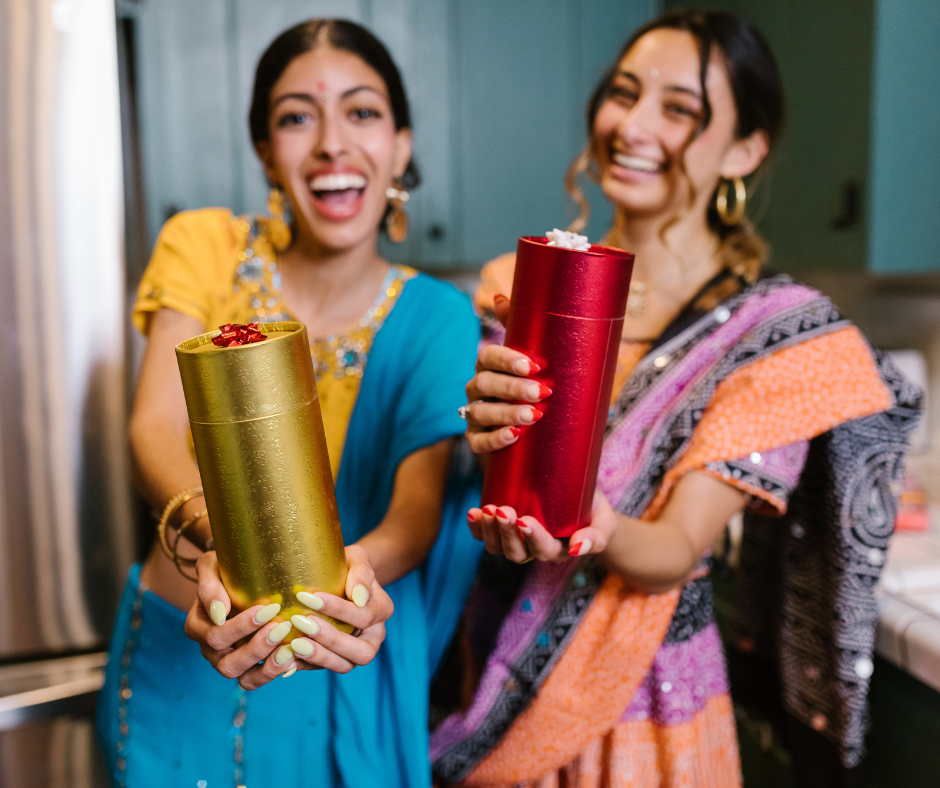 two women wearing traditional kanjeevaram saree holding up a yarn of thread. yarns are of red and gold colour.